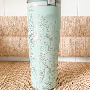 Peut inclure: Une gourde isotherme vert menthe avec un motif floral argenté. La bouteille a un couvercle blanc à bouton-poussoir et une poignée beige. Le motif floral comprend des fleurs d'hibiscus et des feuilles tropicales. La bouteille est conçue pour garder les boissons froides.
