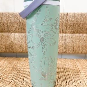 Peut inclure: Un gobelet isotherme vert menthe avec un motif floral argenté. Le gobelet a une poignée lavande et un couvercle blanc. Le motif présente un motif répété de fleurs et de feuilles. Le gobelet est conçu pour garder les boissons froides ou chaudes.