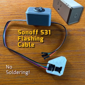 May include: A white Sonoff S31 smart plug with a black power cord and a grey 3D printed box with a black toggle switch. The text "Sonoff S31 Flashing Cable" is written in yellow on a wooden surface. The text "No Soldering!" is written in black on the wooden surface.