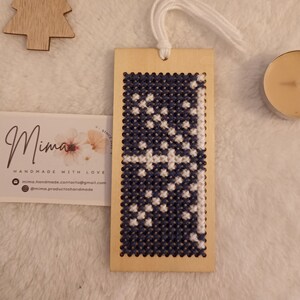 May include: A wooden bookmark with a blue and white cross-stitch design. The bookmark has a white tassel and is handmade. The text "Mima Handmade with Love" is on a card in the image.