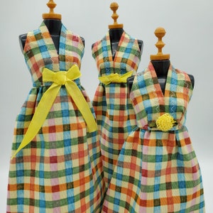 May include: Three doll dresses in a plaid pattern with yellow sashes and a yellow flower accent. The dresses are in shades of blue, green, orange, and pink.