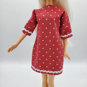 May include: A red doll dress with white polka dots and white trim. The dress has short sleeves and a simple, classic design. The doll's blonde hair is visible.