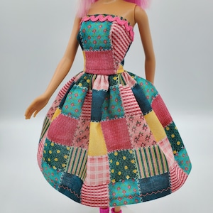 May include: A doll wearing a strapless patchwork dress. The dress features a mix of floral, striped, and solid fabric squares in teal, pink, yellow, and navy blue. The bodice has a pink scalloped trim, and the skirt is full and flared.