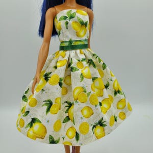 May include: A doll dress featuring a white fabric base with a repeating pattern of yellow lemons and green leaves. The dress has a fitted bodice and a flared skirt, accented by a solid green horizontal band at the waist. The doll has dark blue hair.