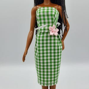 May include: A doll wearing a strapless, knee-length dress featuring a green and white gingham check pattern. The dress is accented with a small, light pink fabric flower attached at the waistline, and the doll has long, dark, braided hair.