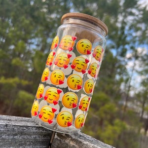 May include: A clear glass water bottle with a bamboo lid. The bottle is decorated with a repeating pattern of yellow emoji faces with red hearts and winking eyes.