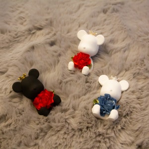 May include: Three bear-shaped figurines with crowns and roses. One black bear with a red rose, and two white bears, one with a red rose and the other with a blue rose. The figurines are on a grey, fluffy surface.