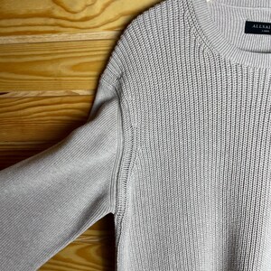 May include: A light beige knitted sweater with a ribbed texture. The sweater has a crew neck and a label that reads "ALLSAINTS" on the bottom right side.