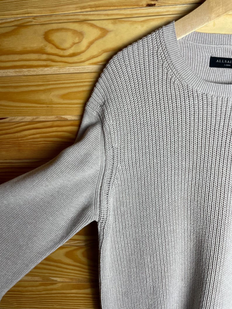 May include: A light beige knitted sweater with a ribbed texture. The sweater has a crew neck and a label that reads "ALLSAINTS" on the bottom right side.