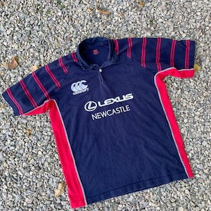 Peut inclure: Un maillot de rugby bleu marine &agrave; rayures rouges et blanches. Le maillot porte le logo Canterbury et un logo Lexus Newcastle.