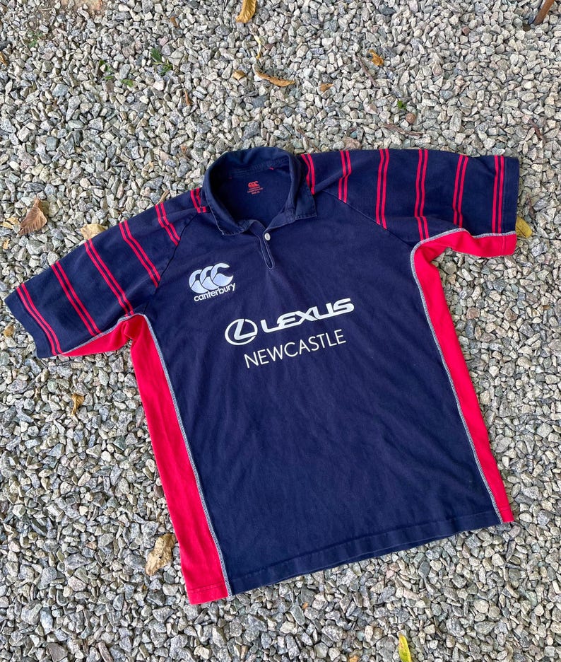 Peut inclure: Un maillot de rugby bleu marine &agrave; rayures rouges et blanches. Le maillot porte le logo Canterbury et un logo Lexus Newcastle.