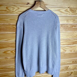 May include: A light gray long-sleeved sweater with a ribbed texture. The sweater has a crew neck and a slightly fitted silhouette.