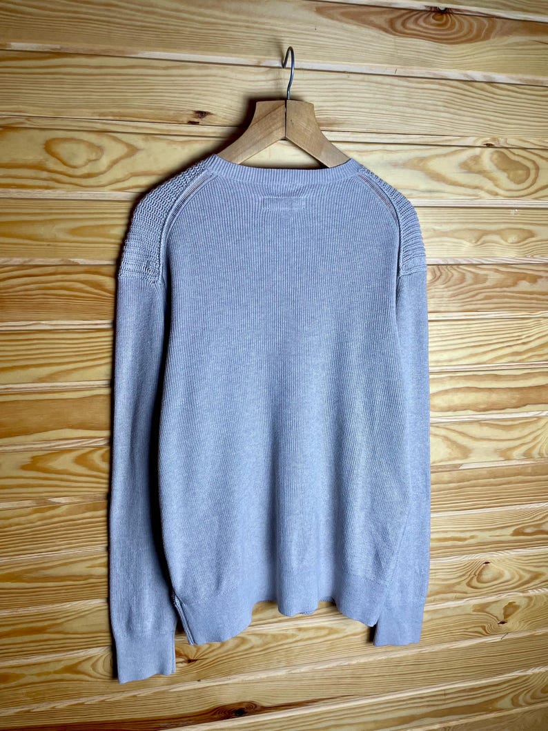 May include: A light gray long-sleeved sweater with a ribbed texture. The sweater has a crew neck and a slightly fitted silhouette.