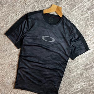 May include: Black athletic t-shirt with a camouflage pattern and the Oakley logo in the center. The shirt has short sleeves and a crew neck. The shirt is hanging on a wooden hanger.