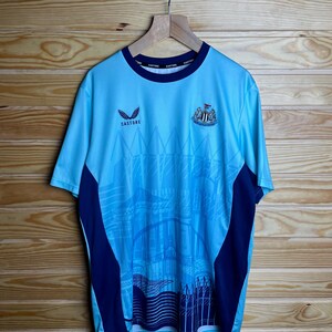 K&ouml;nnte beinhalten: Ein hellblaues Fu&szlig;balltrikot mit dunkelblauem Besatz und einem Teamlogo. Das Trikot zeigt auf der Vorderseite eine Grafik eines Stadions. Der Text "CASTORE" ist auf der Vorderseite des Trikots aufgedruckt.