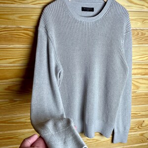 May include: A gray knitted sweater with long sleeves and a crew neck. The sweater has a subtle ribbed texture and the brand name "ALLSAINTS" is visible on the label.