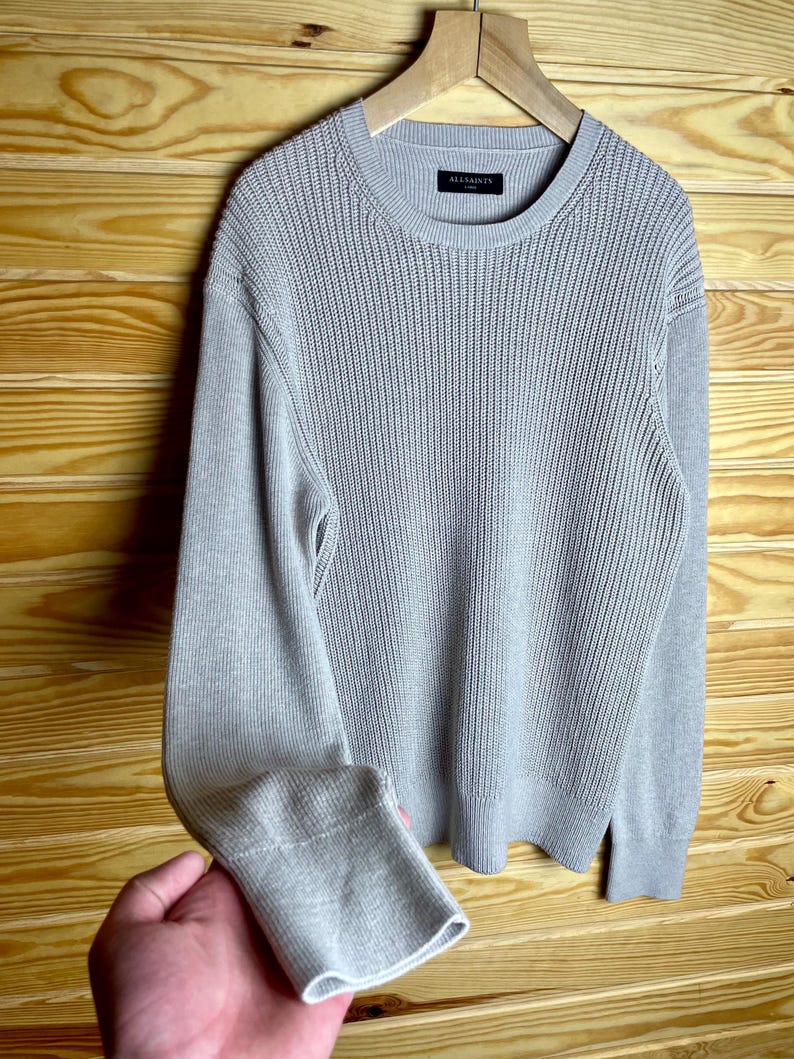May include: A gray knitted sweater with long sleeves and a crew neck. The sweater has a subtle ribbed texture and the brand name "ALLSAINTS" is visible on the label.