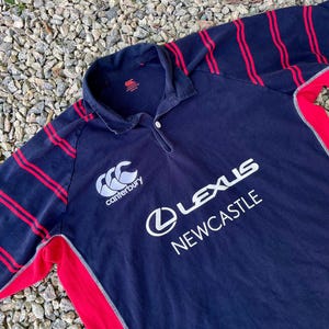 Peut inclure: Un maillot de rugby bleu marine &agrave; rayures rouges et blanches. Le maillot porte le logo Canterbury et le texte "Lexus Newcastle" sur le devant.