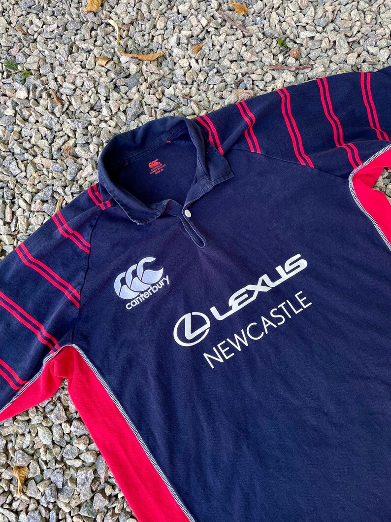 Peut inclure: Un maillot de rugby bleu marine &agrave; rayures rouges et blanches. Le maillot porte le logo Canterbury et le texte "Lexus Newcastle" sur le devant.