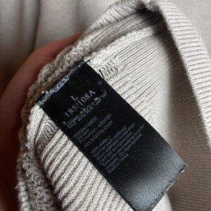 May include: A black tag with white text attached to a light beige sweater. The tag reads "Rothay Crew", "MK029H", "PO number: AS82628R", "ALLSAINTS.COM", "100% cotton", "Machine wash inside out", "Wash separately in cold water", "Cool iron on reverse side", "Take care when wearing to avoid snagging", "Dry clean optional", "Made in China".