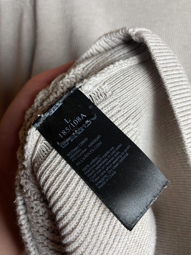 May include: A black tag with white text attached to a light beige sweater. The tag reads "Rothay Crew", "MK029H", "PO number: AS82628R", "ALLSAINTS.COM", "100% cotton", "Machine wash inside out", "Wash separately in cold water", "Cool iron on reverse side", "Take care when wearing to avoid snagging", "Dry clean optional", "Made in China".
