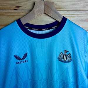 K&ouml;nnte beinhalten: Ein hellblaues Fu&szlig;balltrikot mit einem schwarz-wei&szlig;en Vereinswappen und den Worten "Newcastle United". Das Trikot hat ein wei&szlig;es Logo mit dem Wort "Castore" auf der Vorderseite und dem Wort "Castore" wiederholt am Kragen.