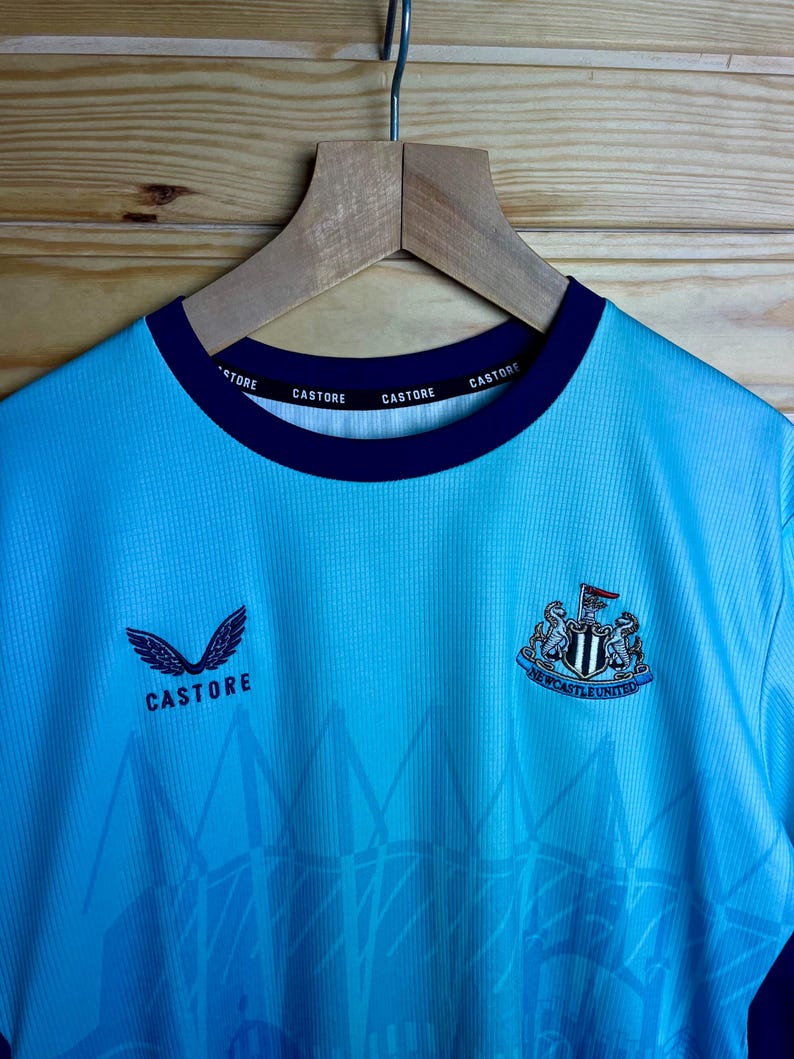 K&ouml;nnte beinhalten: Ein hellblaues Fu&szlig;balltrikot mit einem schwarz-wei&szlig;en Vereinswappen und den Worten "Newcastle United". Das Trikot hat ein wei&szlig;es Logo mit dem Wort "Castore" auf der Vorderseite und dem Wort "Castore" wiederholt am Kragen.