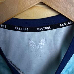 K&ouml;nnte beinhalten: Ein hellblaues Sporttrikot mit marineblauem Besatz. Das Trikot tr&auml;gt den Schriftzug "CASTORE" am Kragen und das Logo der Marke "CASTORE" mit Fl&uuml;geln auf der Vorderseite. Das Gr&ouml;&szlig;enetikett zeigt "2XL".