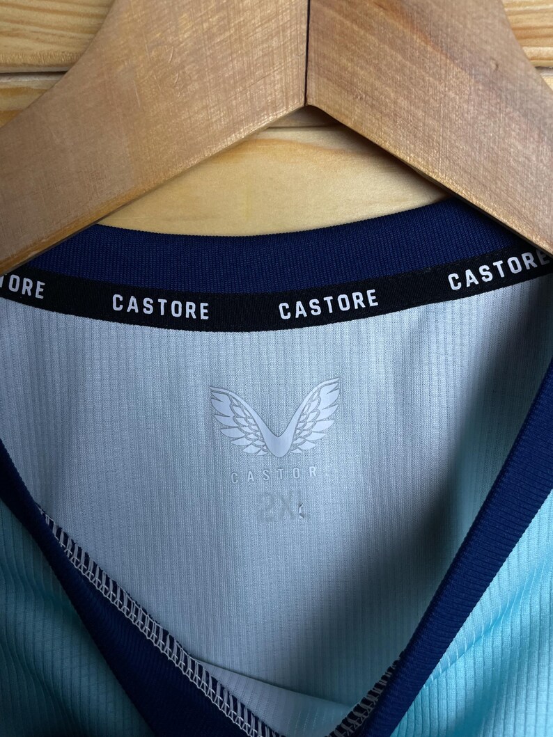 K&ouml;nnte beinhalten: Ein hellblaues Sporttrikot mit marineblauem Besatz. Das Trikot tr&auml;gt den Schriftzug "CASTORE" am Kragen und das Logo der Marke "CASTORE" mit Fl&uuml;geln auf der Vorderseite. Das Gr&ouml;&szlig;enetikett zeigt "2XL".