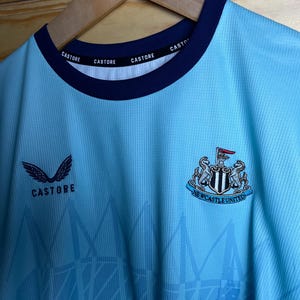 K&ouml;nnte beinhalten: Ein hellblaues Fu&szlig;balltrikot mit einem schwarz-wei&szlig;en Newcastle United Wappen auf der Vorderseite. Das Trikot hat einen dunkelblauen Kragen mit dem Wort "CASTORE" viermal wiederholt. Das Trikot hat auch ein wei&szlig;es "CASTORE" Logo mit Fl&uuml;geln auf der linken Seite der Brust.