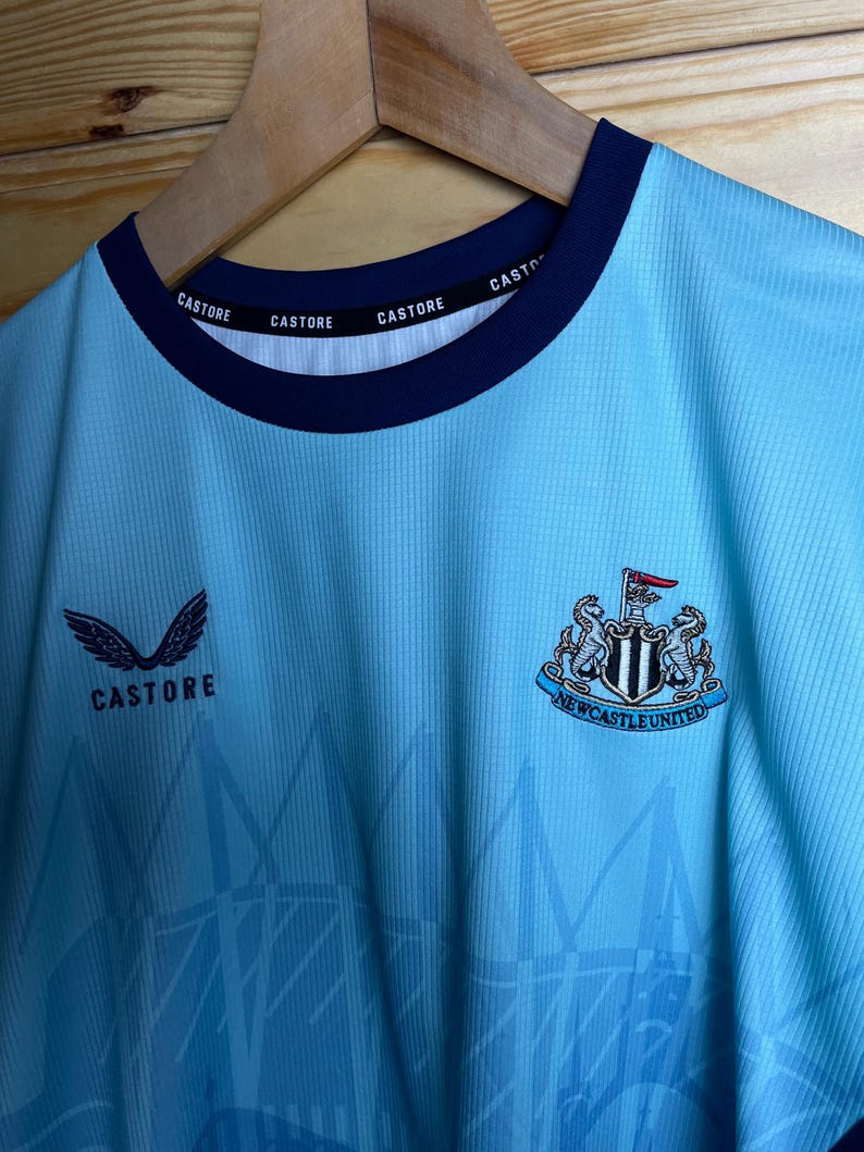K&ouml;nnte beinhalten: Ein hellblaues Fu&szlig;balltrikot mit einem schwarz-wei&szlig;en Newcastle United Wappen auf der Vorderseite. Das Trikot hat einen dunkelblauen Kragen mit dem Wort "CASTORE" viermal wiederholt. Das Trikot hat auch ein wei&szlig;es "CASTORE" Logo mit Fl&uuml;geln auf der linken Seite der Brust.