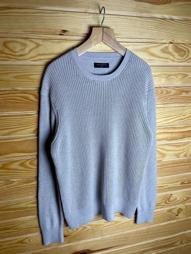 May include: A light gray knitted sweater with long sleeves and a crew neck. The sweater has a small black label with the text "ALLSAINTS" on the front.
