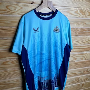 K&ouml;nnte beinhalten: Ein hellblaues Fu&szlig;balltrikot mit dunkelblauem Besatz und einem Teamlogo auf der Vorderseite. Das Trikot hat ein Grafikdesign einer Stadtlandschaft auf der Vorderseite. Der Text "Castore" ist auf der Vorderseite des Trikots aufgedruckt.