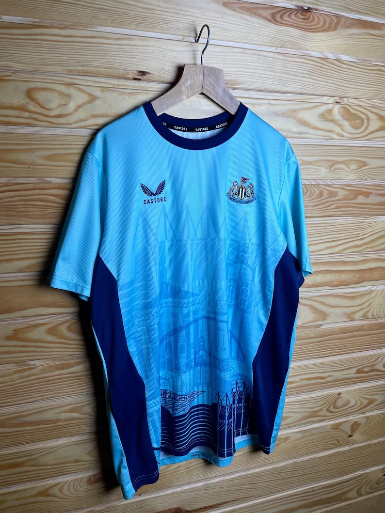 K&ouml;nnte beinhalten: Ein hellblaues Fu&szlig;balltrikot mit dunkelblauem Besatz und einem Teamlogo auf der Vorderseite. Das Trikot hat ein Grafikdesign einer Stadtlandschaft auf der Vorderseite. Der Text "Castore" ist auf der Vorderseite des Trikots aufgedruckt.