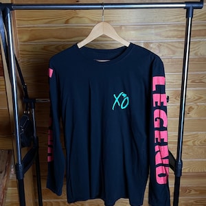 May include: Black long-sleeve shirt with the text "XO" in teal on the chest and "LEGEND" in red on the right sleeve. The shirt is hanging on a wooden hanger.