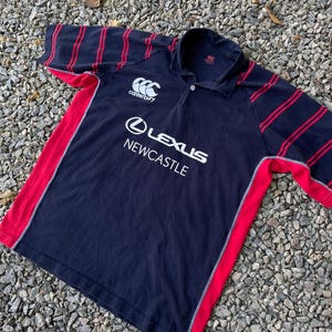 Peut inclure: Un maillot de rugby bleu marine &agrave; rayures rouges et blanches. Le maillot porte le logo Canterbury sur la poitrine gauche et les mots "Lexus Newcastle" sur le devant.