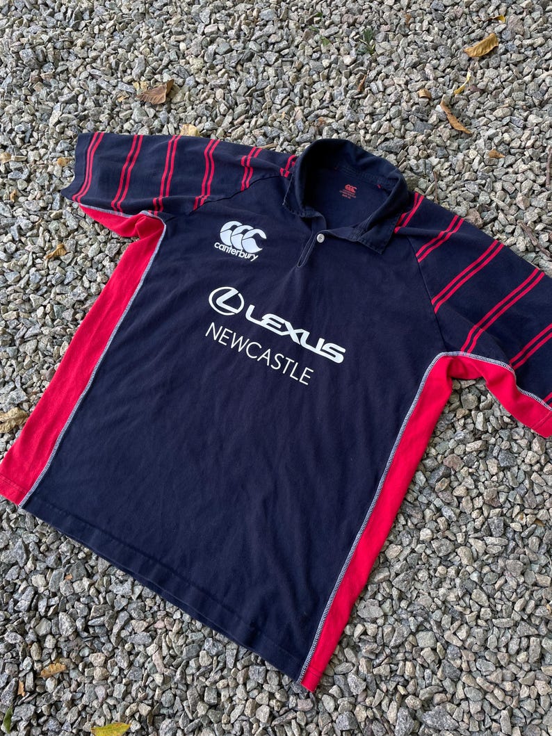 Peut inclure: Un maillot de rugby bleu marine &agrave; rayures rouges et blanches. Le maillot porte le logo Canterbury sur la poitrine gauche et les mots "Lexus Newcastle" sur le devant.