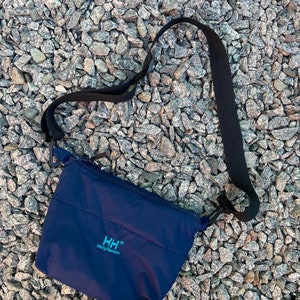 May include: A dark blue crossbody bag with a zipper closure and a black adjustable strap. The bag has a small logo that says "HH® Helly Hansen" in white and light blue lettering.