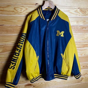 May include: A blue and yellow satin bomber jacket with a large "M" on the chest and "WOLVERINES" on the left sleeve. The jacket has a zipper closure and ribbed cuffs and waistband.
