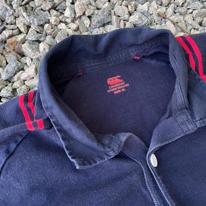 Peut inclure: Un maillot de rugby noir avec des rayures rouges et blanches sur les manches. Le maillot a un logo blanc Canterbury of New Zealand sur le devant et une &eacute;tiquette de taille indiquant "Taille XL".