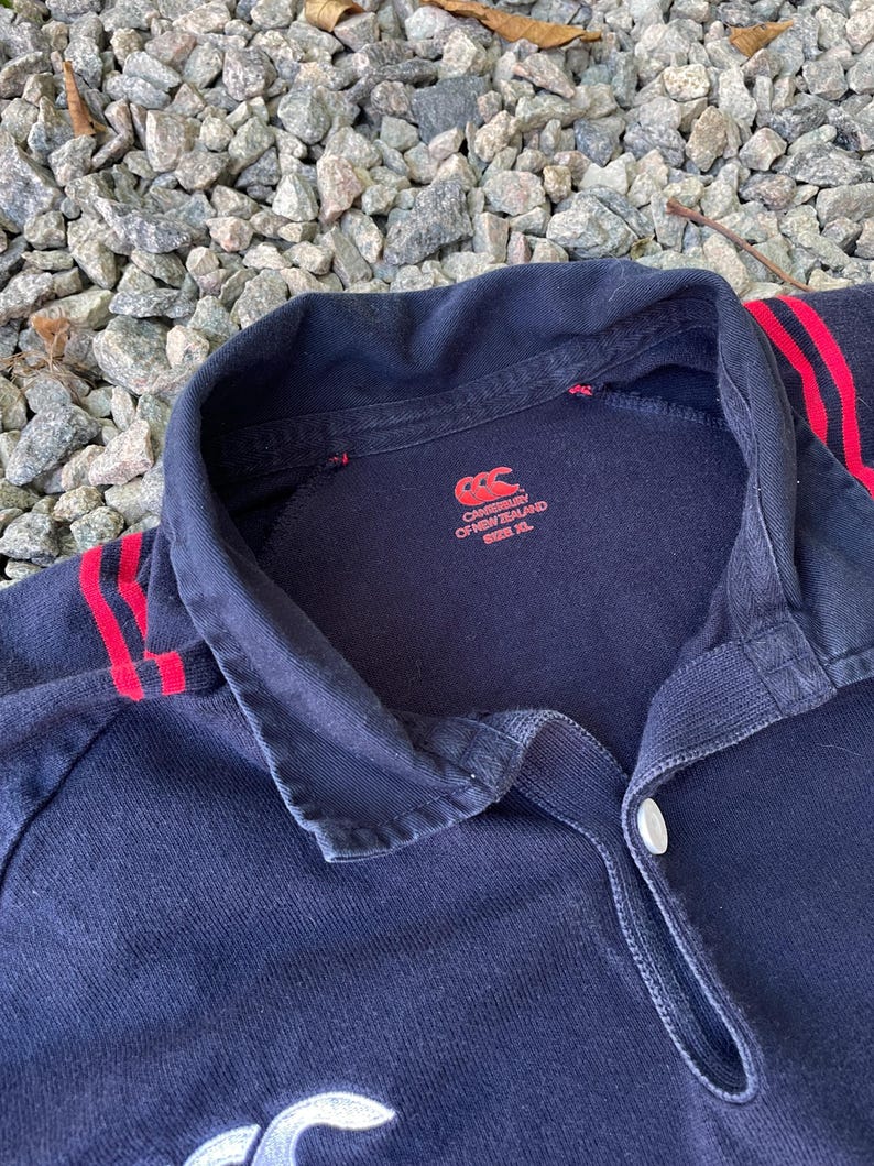 Peut inclure: Un maillot de rugby noir avec des rayures rouges et blanches sur les manches. Le maillot a un logo blanc Canterbury of New Zealand sur le devant et une &eacute;tiquette de taille indiquant "Taille XL".