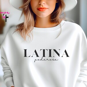 May include: A white sweatshirt featuring the word "LATINA" in a bold, black font, with "poderosa" in a smaller, cursive font beneath. The sweatshirt has long sleeves and a crew neck. The image also includes the text "white" in the bottom right corner.