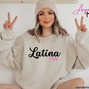 May include: Sandstone-colored sweatshirt with the word "Latina" in black script and "PWR" in pink. The sweatshirt is worn by a person making a peace sign with their fingers. The words "Amor y Fe" are in pink script.