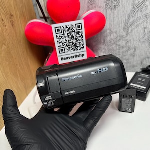 Puede incluir: Una videocámara negra Panasonic HC-V760 Full HD con capacidad de zoom de 50x. El dispositivo cuenta con un control de lente lateral y un botón de función de cámara. Se incluyen una batería negra separada y un adaptador de corriente negro con cable.
