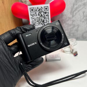 May include: A black Sony digital camera featuring a 20x optical zoom lens, a wrist strap, a rechargeable battery pack, and a white USB charging cable. The camera body displays the Sony brand name and lens specifications on the front.