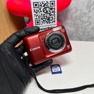 May include: A red Canon digital camera with a black strap and a 4GB memory card. The camera has a silver lens and the text "Canon" is visible on the front. The camera has a 3.3x optical zoom and 10.0 megapixels.