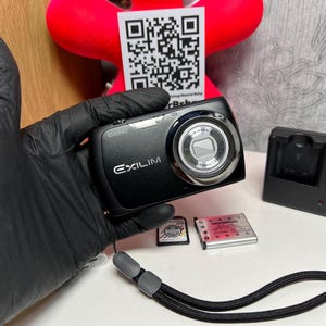 Pode incluir: Câmera digital Casio Exilim preta com lente prateada, segurada por uma mão enluvada. Inclui uma alça de pulso, um carregador de bateria e dois cartões de memória. A câmera tem a marca "Exilim" impressa.