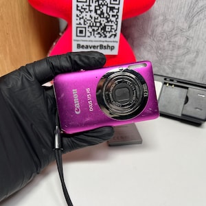 May include: A magenta Canon IXUS 115 HS digital camera with a silver lens and a black wrist strap. The camera is held in a gloved hand. A black battery charger is in the background.