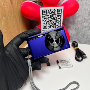 Puede incluir: Una cámara digital Lumix de color azul real con un objetivo negro, que incluye una correa de muñeca gris, un cable de carga USB negro, una batería rectangular y una pequeña tarjeta de memoria. Una tarjeta blanca con un código QR negro al fondo.