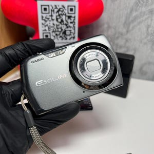 May include: A silver Casio Exilim digital camera featuring a prominent circular lens housing with text reading 4X OPTICAL ZOOM and f=4.9-19.6mm 1:3.2-6.5. The compact device includes a wrist strap and displays the brand name Casio and model name Exilim.